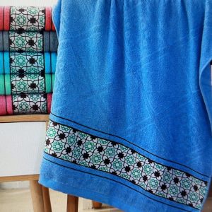 Blue Moroccan style geometric bath towel on wooden chair with black-white-green border, matching towels in coral, grey, teal, green, and pink stacked on cabinet